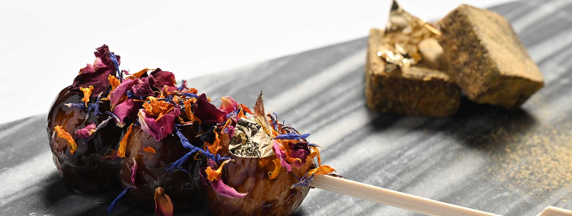Brochette de dessert décorée de fleurs comestibles, présentée sur une ardoise avec des cubes dorés en arrière-plan.